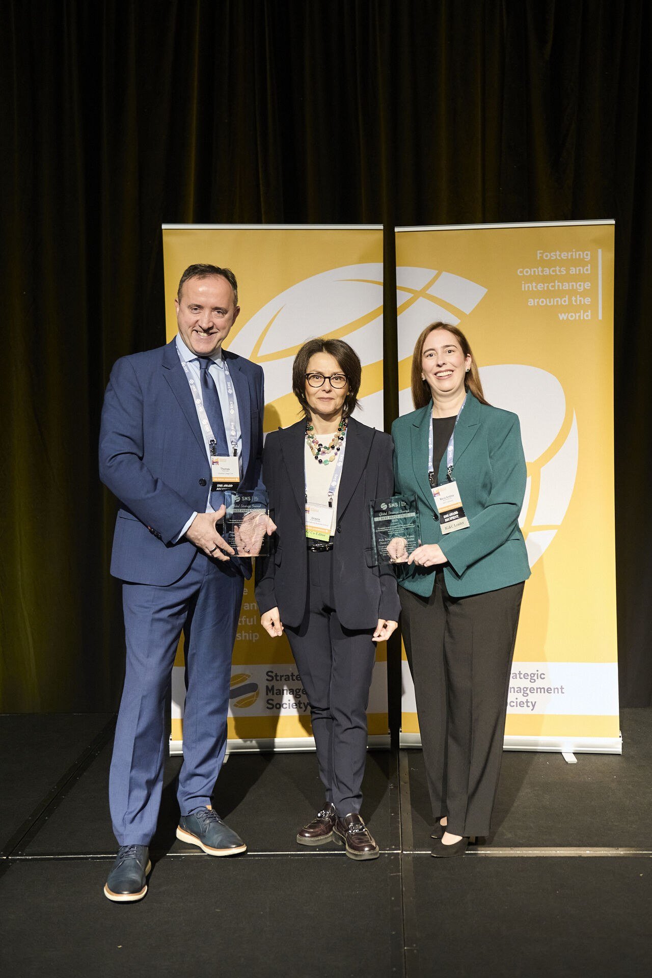 Thomas C. Lawton, left, with two colleagues on stage at a Strategic Management Society awards ceremony, each holding a clear award plaque in front of yellow SMS banners.